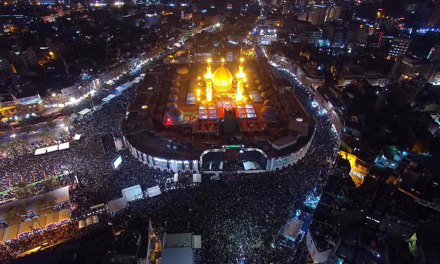 Aerial night view of the illuminated golden dome and minarets of the Shrine of Imam Hussain (AS), surrounded by crowds of pilgrims