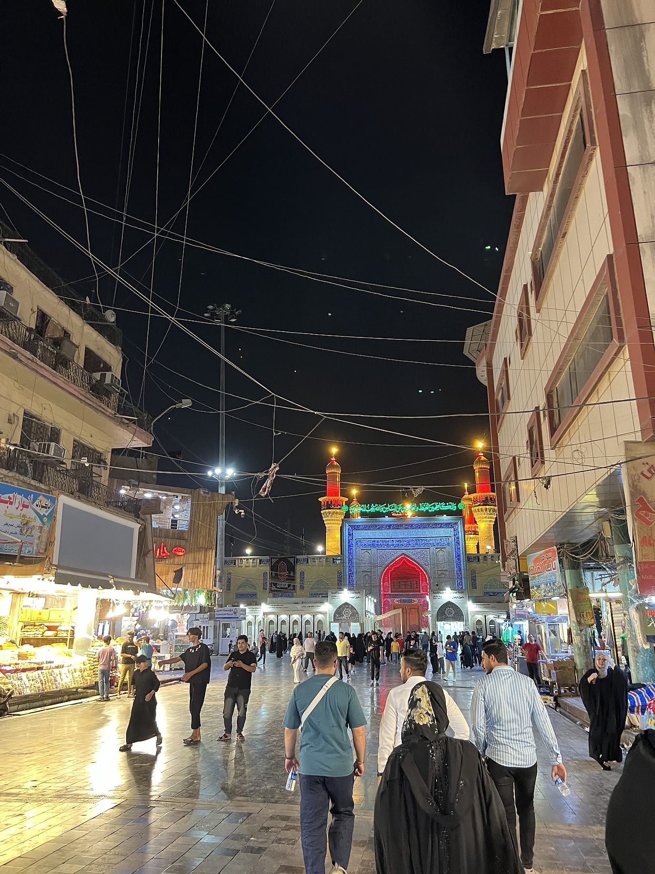 A busy lit street lined with shops at night, leading towards a shrine gateway