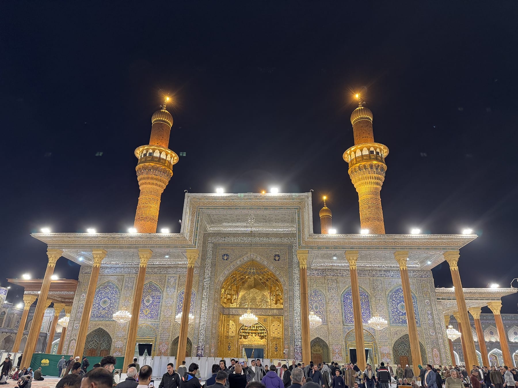 The illuminated shrine façade at night, with a tiled golden entrance arch between two tall gold minarets