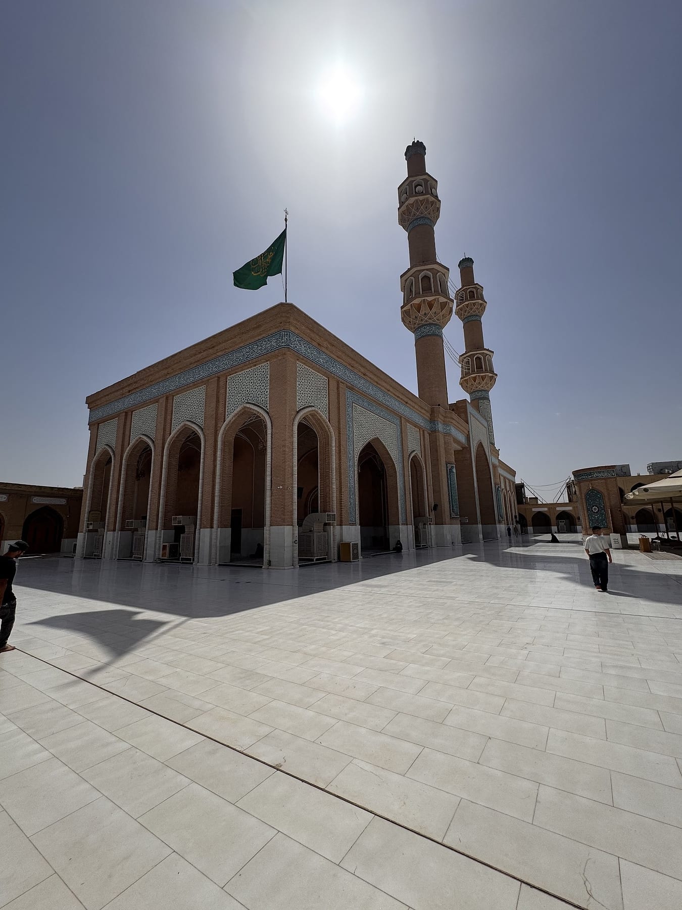 A brick arcade with a green flag on the roof and two tall minarets, under a bright sky