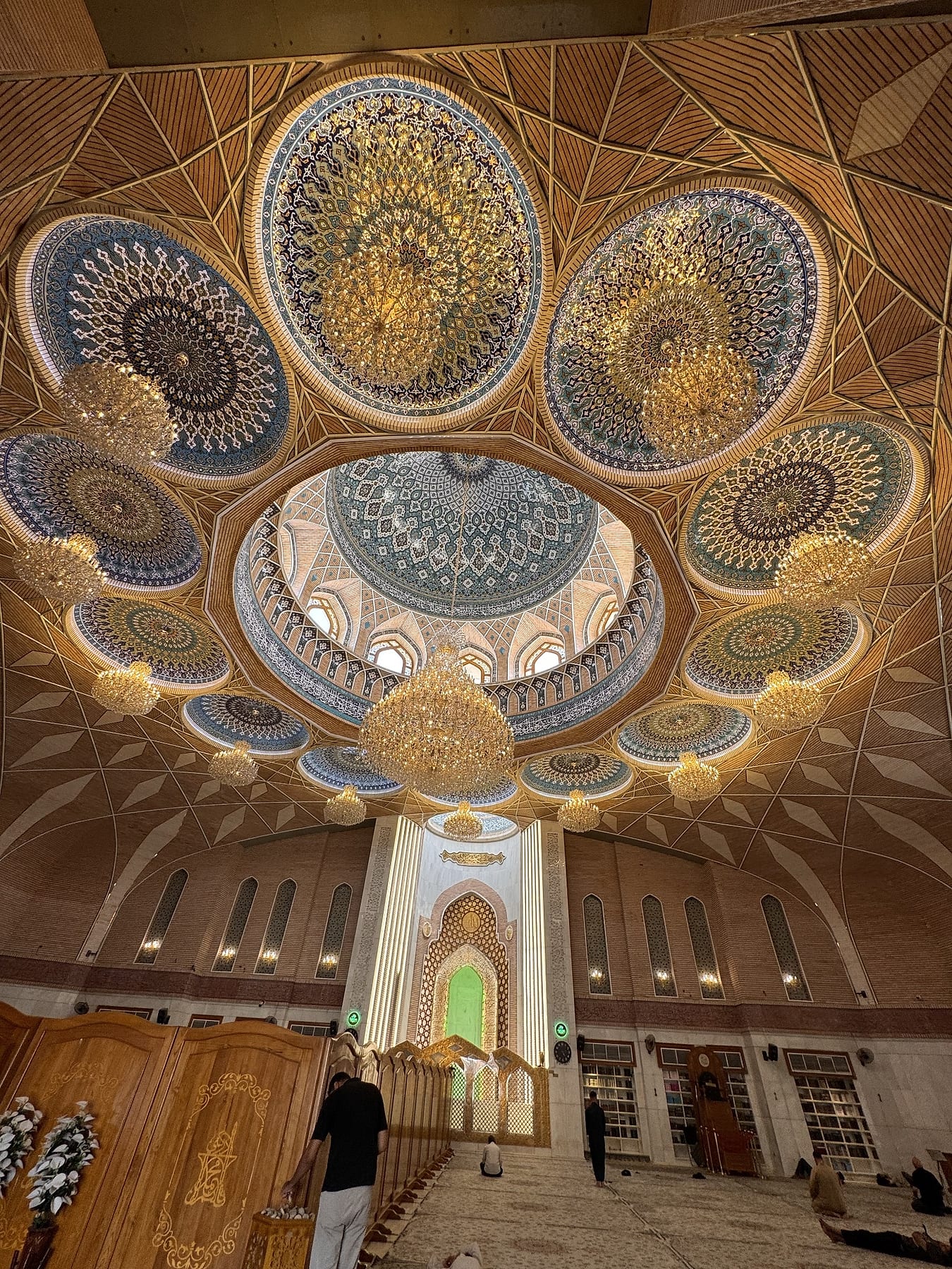 A richly painted domed ceiling of blue and gold medallions with chandeliers, above a lit prayer niche