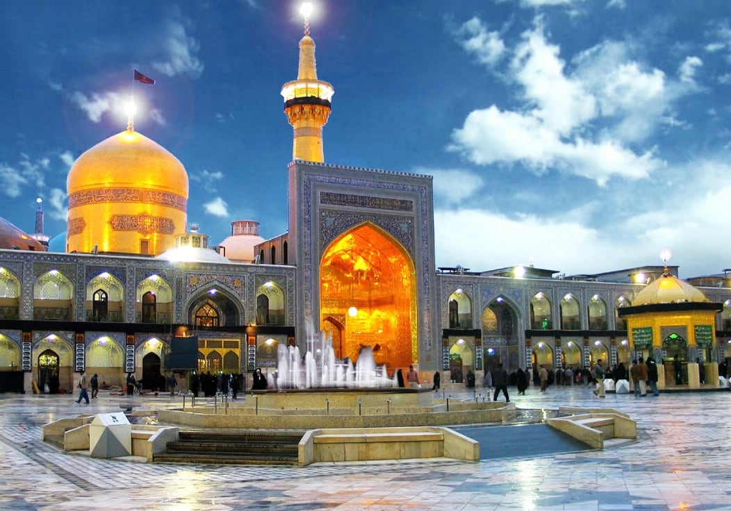 A golden dome and a golden minaret above a tiled courtyard with a glowing golden iwan, a fountain and visitors, under a blue evening sky