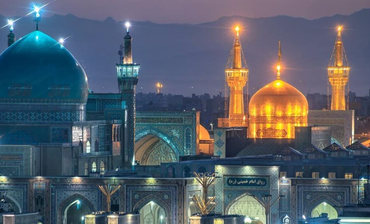 The shrine complex at night: a large turquoise dome on the left, and the golden dome between two lit golden minarets, with mountains behind