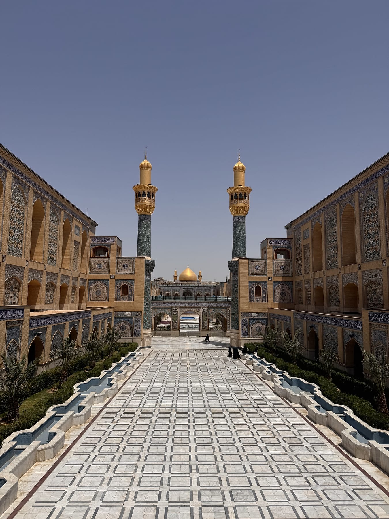 A long tiled walkway between arcaded courtyard buildings leads towards a golden dome flanked by two minarets