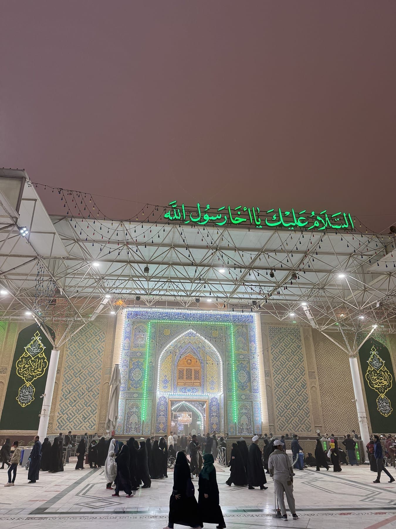 A brightly lit entrance at dusk, with green Arabic lettering above it and visitors in the courtyard
