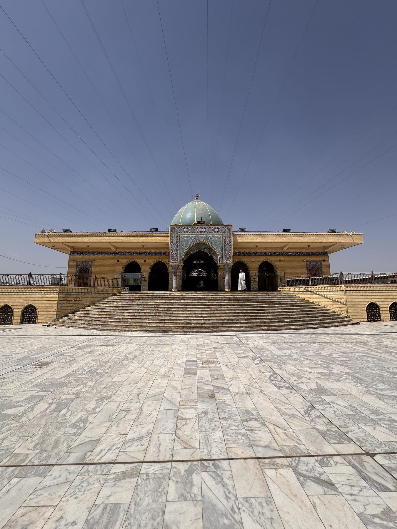A low arcaded building with a small turquoise dome, above a wide stairway and a marble forecourt